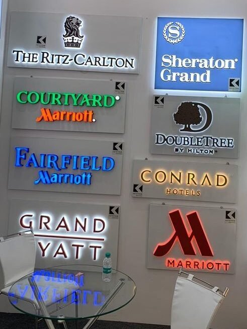 "Indoor display of various illuminated hotel signage boards showcasing different styles of LED, acrylic, and backlit signage designs, arranged on a showroom wall for commercial branding purposes".