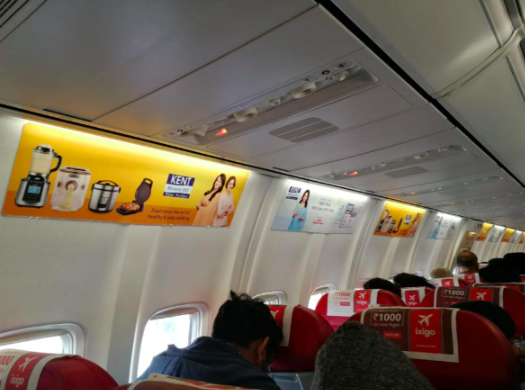 Inflight advertisements displayed above passenger windows in an airplane, featuring product ads from brands like Kent and Big Basket.